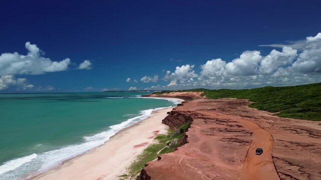 Aerial view of a vehicle driving on a red dirt road along the cliffs overlooking the turquoise ocean at Pipa Beach, Rio Grande do Norte, Brazil.