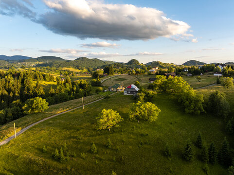 Aerial view of the rural landscape featuring rolling green hills, scattered houses, lush trees, and a winding road under a blue sky with soft clouds in Sirnea, Brasov, Romania.