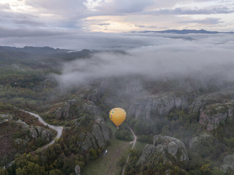 Aerial view of a yellow hot air balloon floating above the dramatic Belogradchik Rocks surrounded by low-hanging clouds and autumn forest in Belogradchik, Vidin, Bulgaria.