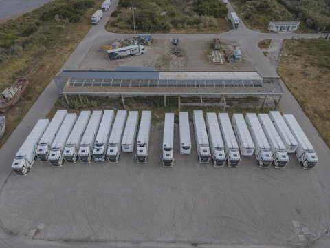 Aerial view of a fleet of white semi-trucks and trailers parked in an orderly row on a gravel lot in Vila do Conde, Porto District, Portugal.