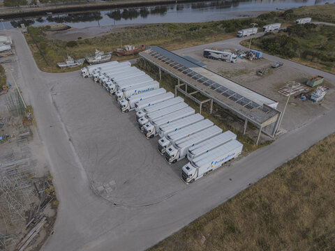 Aerial view of a fleet of white Primafrio semi-trucks parked in a row on a paved lot near an estuary with old boats in Vila do Conde, Porto District, Portugal.