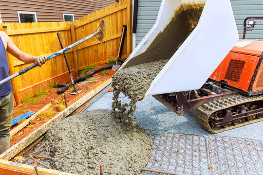 Worker uses tracked dumper mixer to pour concrete into framework for patio in backyard area.