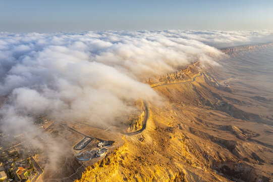 Aerial view of Mitzpe Ramon crater with a thick layer of clouds spilling over the edge of the desert cliffs, illuminated by golden hour sunlight in Mitzpe Ramon, South District, Israel.