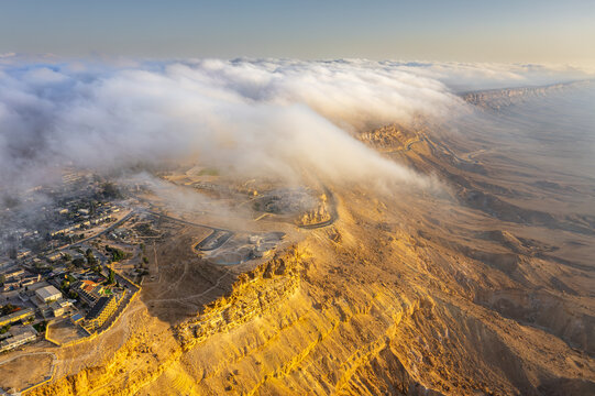 Aerial view of the Makhtesh Ramon crater with low-hanging clouds spilling over the rugged desert cliffs at golden hour in Mitzpe Ramon, South District, Israel.