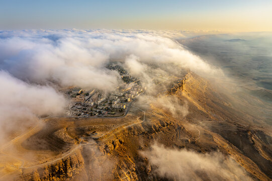 Aerial view of Mitzpe Ramon crater partially covered by low-hanging clouds at sunset with desert cliffs and town buildings in Mitzpe Ramon, South District, Israel.