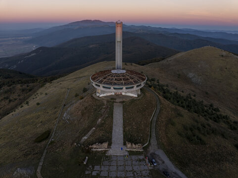 Aerial view of Buzludzha Monument, a brutalist structure atop a mountain peak during golden hour with rolling hills and distant mountains, Stara Zagora, Bulgaria.