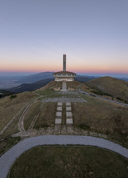 Aerial view of the Buzludzha Monument, a brutalist structure on a mountain peak at sunset, with paved paths and grassy slopes in Stara Zagora, Bulgaria.