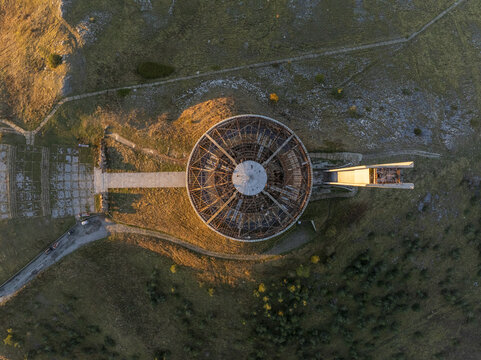 Aerial view of the Buzludzha Monument, a circular brutalist structure with exposed framework, set against the grassy mountain landscape during golden hour in Stara Zagora, Bulgaria.