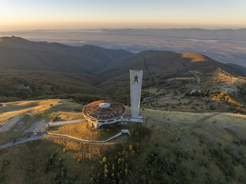Aerial view of the iconic Buzludzha Monument, featuring its saucer-shaped structure and tower with a red star, situated atop a mountain peak during golden hour in Stara Zagora, Bulgaria.