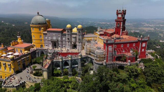 Aerial view of the colorful Pena Palace with its distinctive red and yellow towers, domes, and surrounding lush forest landscape in Sintra, Portugal.