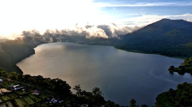 Aerial view of Danau Buyan surrounded by lush green hills and mist under the golden hour sun in Sukasada, Bali, Indonesia.