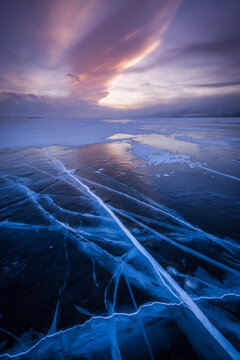 View of the frozen surface of Lake Baikal with intricate ice cracks and patterns under a dramatic sunset sky in Irkutsk, Irkutsk Oblast, Russia.