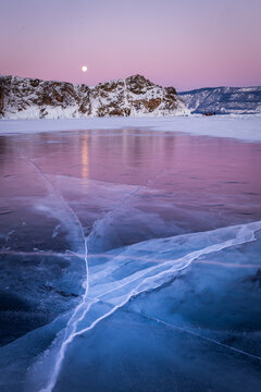 View of the frozen surface of Lake Baikal with deep ice cracks under a soft pink sunset sky and a visible moon above the snowy mountains in Irkutsk, Irkutsk Oblast, Russia.