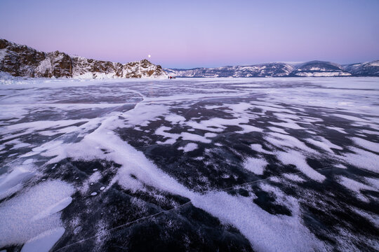 View of the frozen surface of Lake Baikal with intricate ice patterns and snow-covered mountains under a twilight sky in Irkutsk, Irkutsk Oblast, Russia.