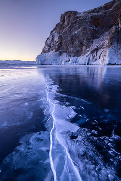 View of Lake Baikal with deep cracks in the frozen ice surface and rocky cliffs in the background during a cold winter sunset in Irkutsk, Irkutsk Oblast, Russia.