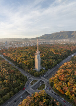 Aerial view of the Sofia TV Tower surrounded by vibrant autumn forest, with a road intersection and tunnel in the foreground, and city skyline in Sofia, Sofia City Province, Bulgaria.