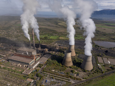 Aerial view of a large coal-fired power plant with massive cooling towers emitting thick plumes of steam into the sky, set within a rural landscape in Bulgaria.