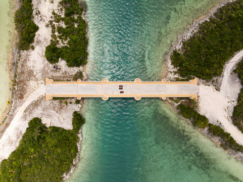 Aerial view of Crab Cay Bridge spanning a narrow turquoise channel surrounded by lush tropical vegetation and white sandy paths in Exuma, The Bahamas.