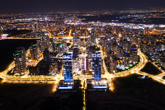 Aerial view of the illuminated skyline at night, featuring modern high-rise residential towers, glowing street lights, and urban infrastructure in Netanya, Center District, Israel.