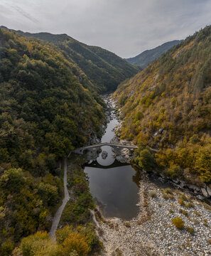 Aerial view of Devil's Bridge spanning the Arda River amidst autumn-colored forests and rocky slopes in Kardzhali Province, Bulgaria.