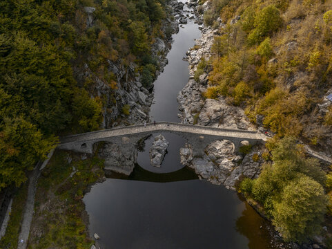Aerial view of the historic stone Devil's bridge spanning the Arda river in a deep rocky gorge surrounded by vibrant autumn forest in Kardzhali Province, Bulgaria.