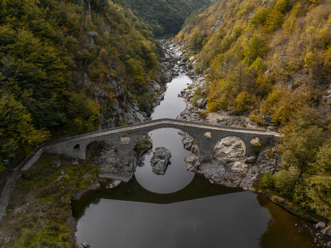Aerial view of Devil's Bridge spanning the Arda River, surrounded by lush autumn forests and rocky terrain in Kardzhali Province, Bulgaria.