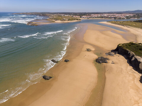 Aerial view of the sandy beach and coastline with gentle waves, rocky outcrops, and the town visible in the distance in Vila Nova de Milfontes, Beja District, Portugal.