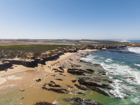 Aerial view of the rugged coastline and sandy beach with dark rock formations, crashing waves, and green coastal vegetation in Vila Nova de Milfontes, Beja District, Portugal.