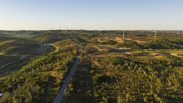 Aerial view of wind turbines situated on rolling green hills with a road winding through the landscape during golden hour in Aljezur, Faro District, Portugal.