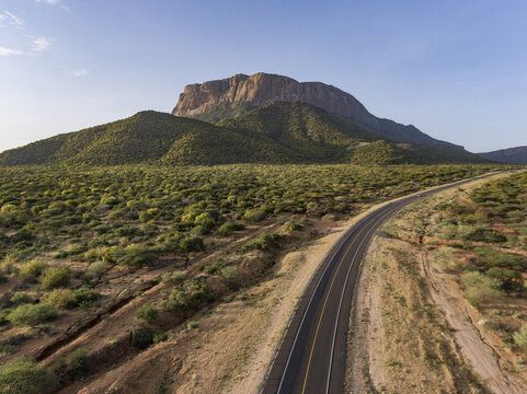 Aerial view of Oldoinyo Sabachi mountain rising above the arid landscape with a winding asphalt road cutting through the sparse vegetation in Archers Post, Samburu County, Kenya.