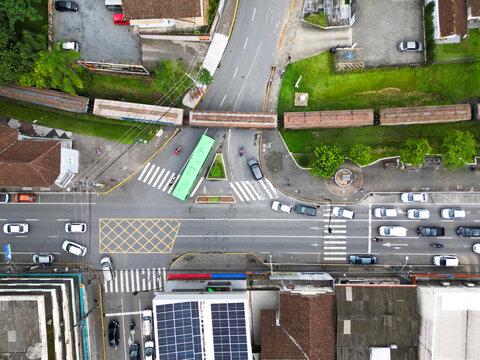 Aerial view of a busy urban intersection with a train crossing the tracks, surrounded by buildings, traffic, and solar panels in Jaragua do Sul, Santa Catarina, Brazil.