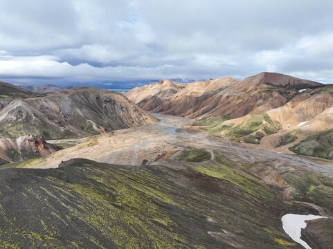 Aerial view of the colorful rhyolite mountains and braided river systems of Landmannalaugar, with mossy slopes and snow patches under a cloudy sky in Hella, Rangarthing ytra, Iceland.