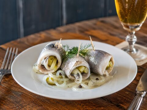 Rollmops im deutschen Restaurant: Eingelegte Heringsfilets mit Gew&uuml;rzgurke und Zwiebeln, auf wei&szlig;em Teller mit Dill angerichtet und passend zu einem k&uuml;hlen Bier serviert.