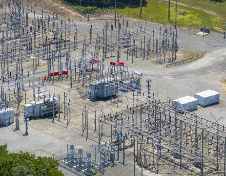 Aerial view of an electrical substation with large transformers, metal power grid structures, and gravel grounds in Commack, New York, United States.