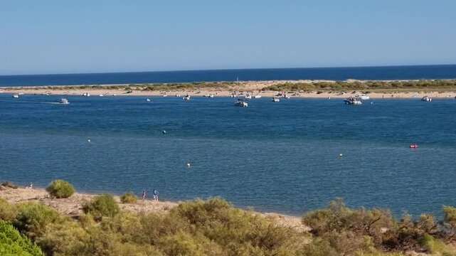 Wide clip of a small ferry and boats cruising the Piedra River mouth near Flecha del Rompido sandbar in Cartaya, Huelva, Spain, under a clear blue sky.