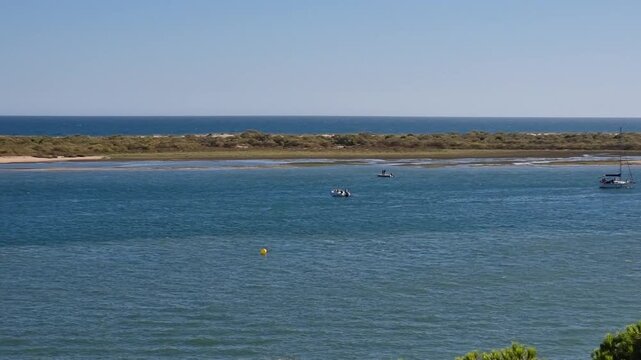 Wide clip of a small ferry and boats cruising the Piedra River mouth near Flecha del Rompido sandbar in Cartaya, Huelva, Spain, under a clear blue sky.