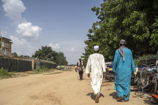 Maroua, Far North Region, Cameroon - 27 September 2021: View of two men in traditional robes walking along a dirt road with parked motorcycles and trees under a blue sky.