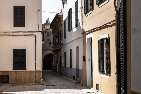 View of a narrow cobblestone street with traditional Mediterranean architecture, white and ochre facades, and dark green shutters in Ciutadella, Balearic Islands, Spain.
