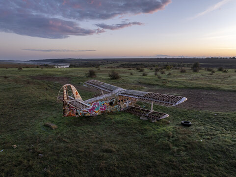 Aerial view of an abandoned, graffiti-covered biplane resting in a grassy field under a soft sunset sky in Bulgaria.