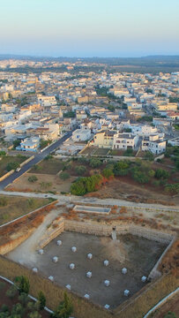 Aerial view of the town of Giuliano Di Lecce with white buildings, olive groves, and a stone-walled excavation site in the foreground in Giuliano Di Lecce, Puglia, Italy.