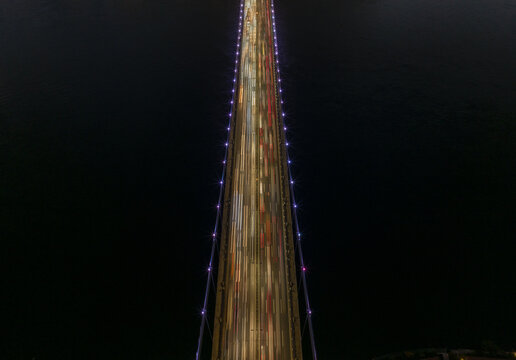 Aerial view of Fatih Sultan Mehmet Bridge at night with long exposure light trails from traffic, illuminated suspension cables, and dark waters in Beykoz, Istanbul, Turkey.
