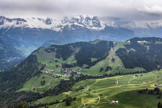 Aerial view of Stanserhorn Cabrio mountain landscape with lush green alpine meadows, winding paths, farmhouses, and snow-capped peaks under a cloudy sky in Stans, Nidwalden, Switzerland.