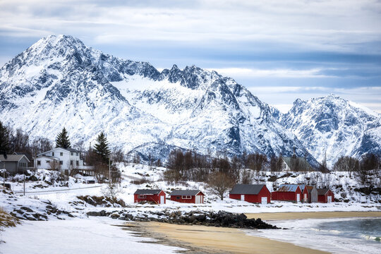 View of the snowy landscape of Senja with traditional red cabins along the sandy beach, backed by dramatic snow-covered mountains under a cloudy sky in Senja, Troms, Norway.