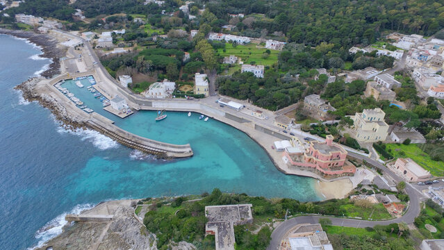 Aerial view of the harbor of Tricase Porto featuring turquoise water, small boats, a historic pink building, and coastal roads in Tricase Porto, Puglia, Italy.
