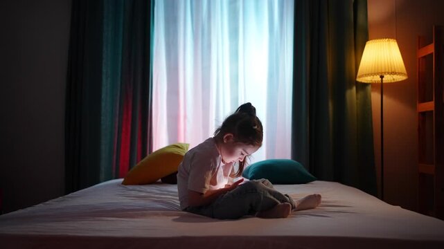 A lonely child sitting on a bed in a dark room in the evening, using a smartphone.