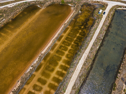 Aerial view of the salt pans in Quelfes featuring rectangular basins with varying water colors, a dirt path, and surrounding marshland in Quelfes, Faro District, Portugal.