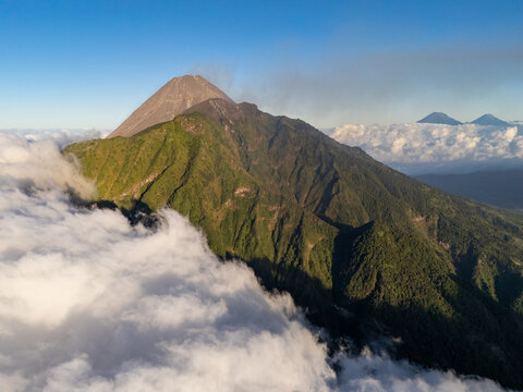 Aerial view of Mount Merapi with its volcanic peak rising above lush green slopes and a sea of clouds in Selo, Central Java, Indonesia.