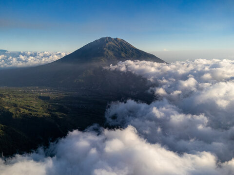 Aerial view of Mount Merbabu rising above a dense sea of clouds with lush green slopes under a clear blue sky in Selo, Central Java, Indonesia.