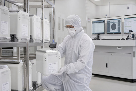 Technician in full cleanroom protective clothing scans a labeled material container inside a modern semiconductor manufacturing facility.