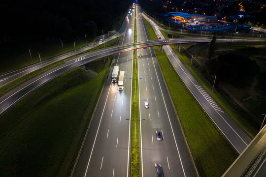 Aerial view of a highway interchange at night with illuminated roads, moving trucks and cars, and green grass verges in Krakow, Lesser Poland Voivode, Poland.